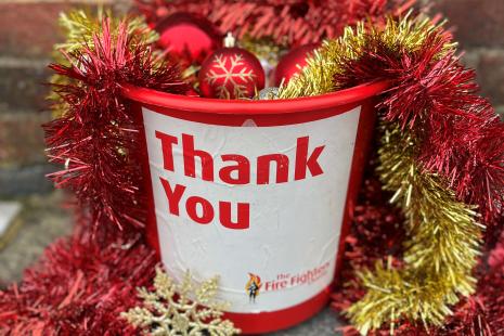 Red bucket in front of brick will on pavement. Words Thank you printed on and The Fire Fighters Charity. Bucket has red and gold tinsel and baubles overflowing from it.
